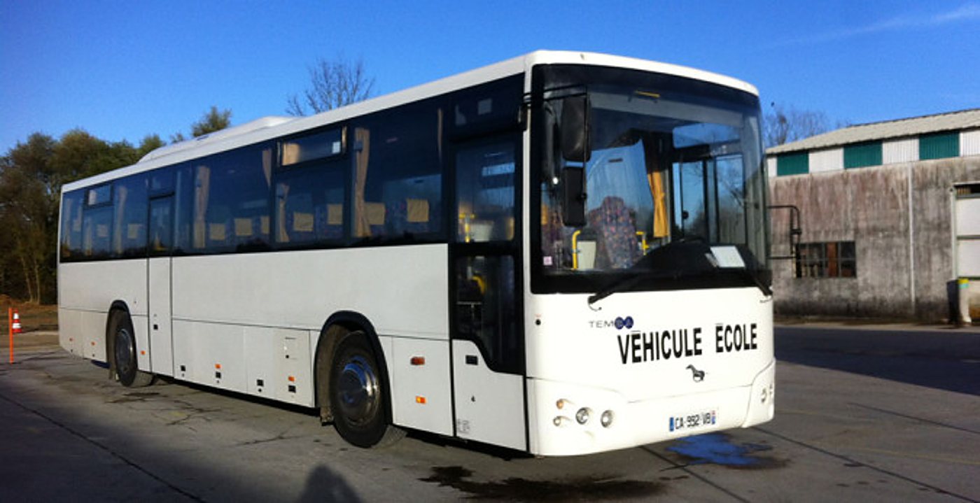 Autocar blanc « Véhicule école » garé devant un bâtiment industriel, sous un ciel bleu.