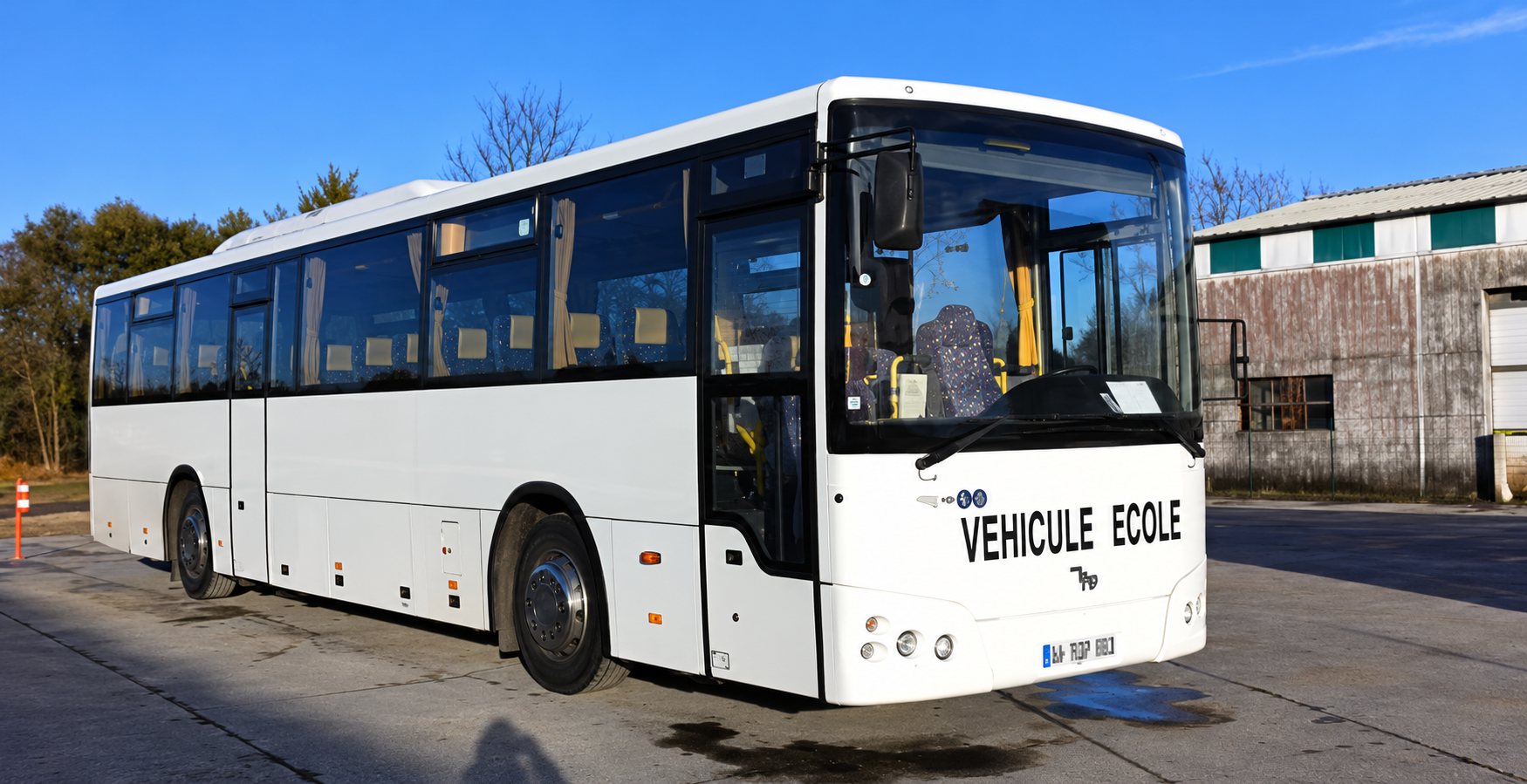 Autocar blanc « Véhicule école » garé sur un parking goudronné, devant des arbres et un bâtiment.