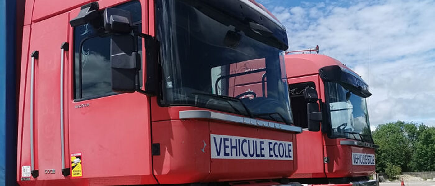 Deux cabines de camions rouges portant l’inscription « Véhicule école », garées côte à côte sous un ciel nuageux.