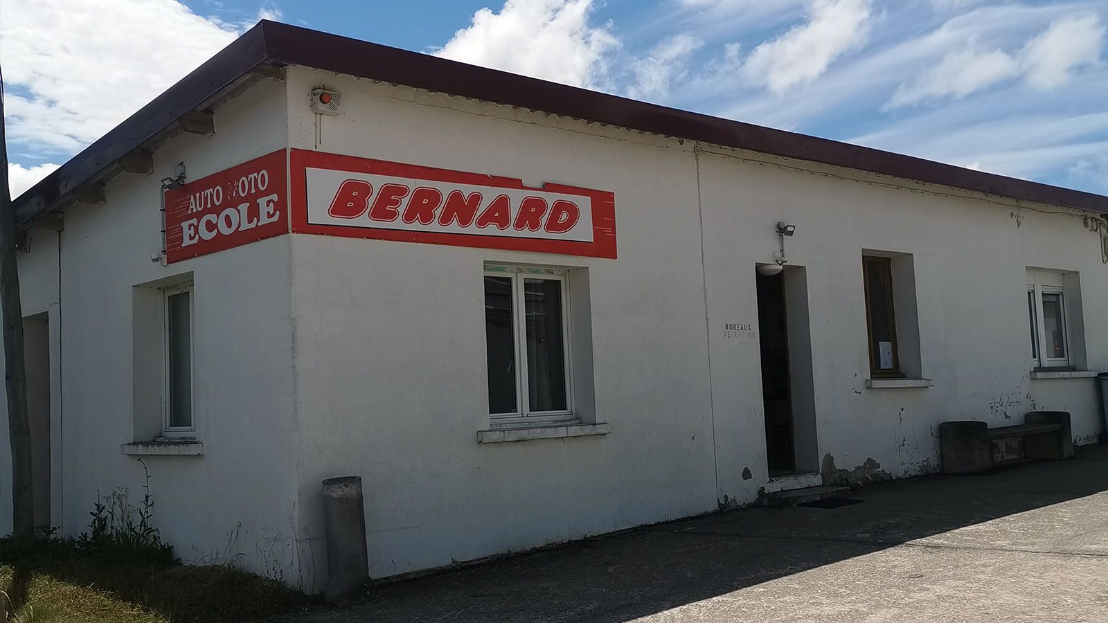 Bâtiment bas aux murs blancs portant l’enseigne rouge Auto Moto École Bernard, sous un ciel bleu nuageux.