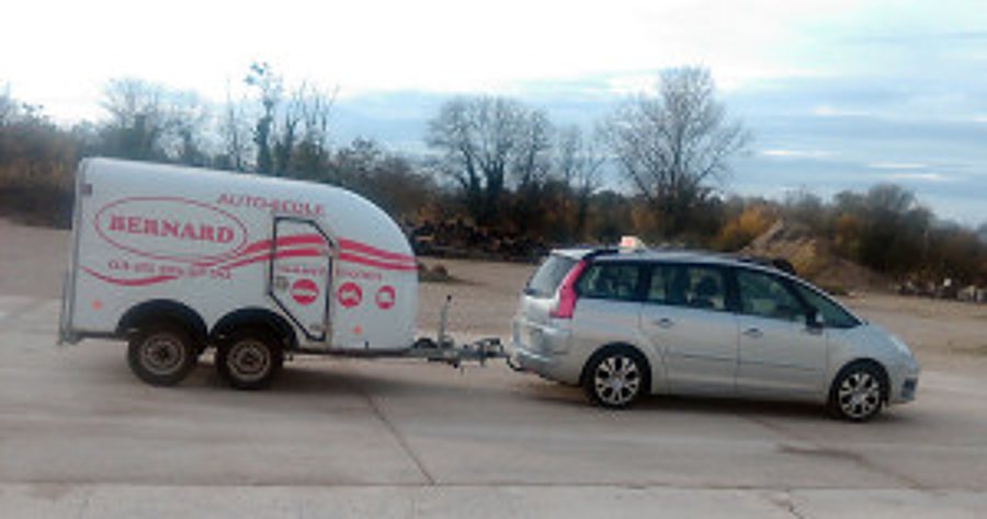 Monospace gris attelé à une remorque blanche portant le nom Bernard, sur un grand parking.